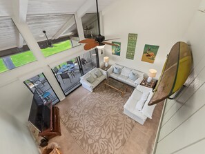 A spacious living area with a white sofa, a coffee table, and a ceiling fan. There is a view of a green lawn through large windows. - Sunset, Ocean, and Golf Course Views! (Waikoloa Village)