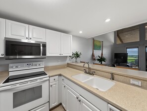 A modern kitchen with white cabinetry, a stainless steel microwave, an oven, a double sink, and a television. - Sunset, Ocean, and Golf Course Views! (Waikoloa Village)