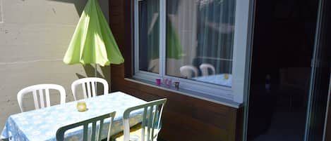 Um pátio com mesa e cadeiras, um guarda-sol verde e uma janela com cortinas.