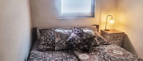 Un dormitorio con cama, almohadas, una mesita de noche con lámpara y una ventana con persianas.