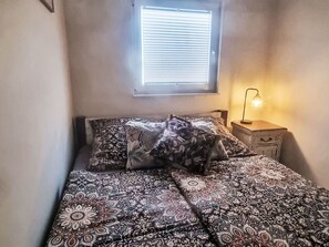 A bedroom with a bed, pillows, a bedside table with a lamp, and a window with blinds. - Gästehaus am Neetzekanal by Interhome (Bardowick)