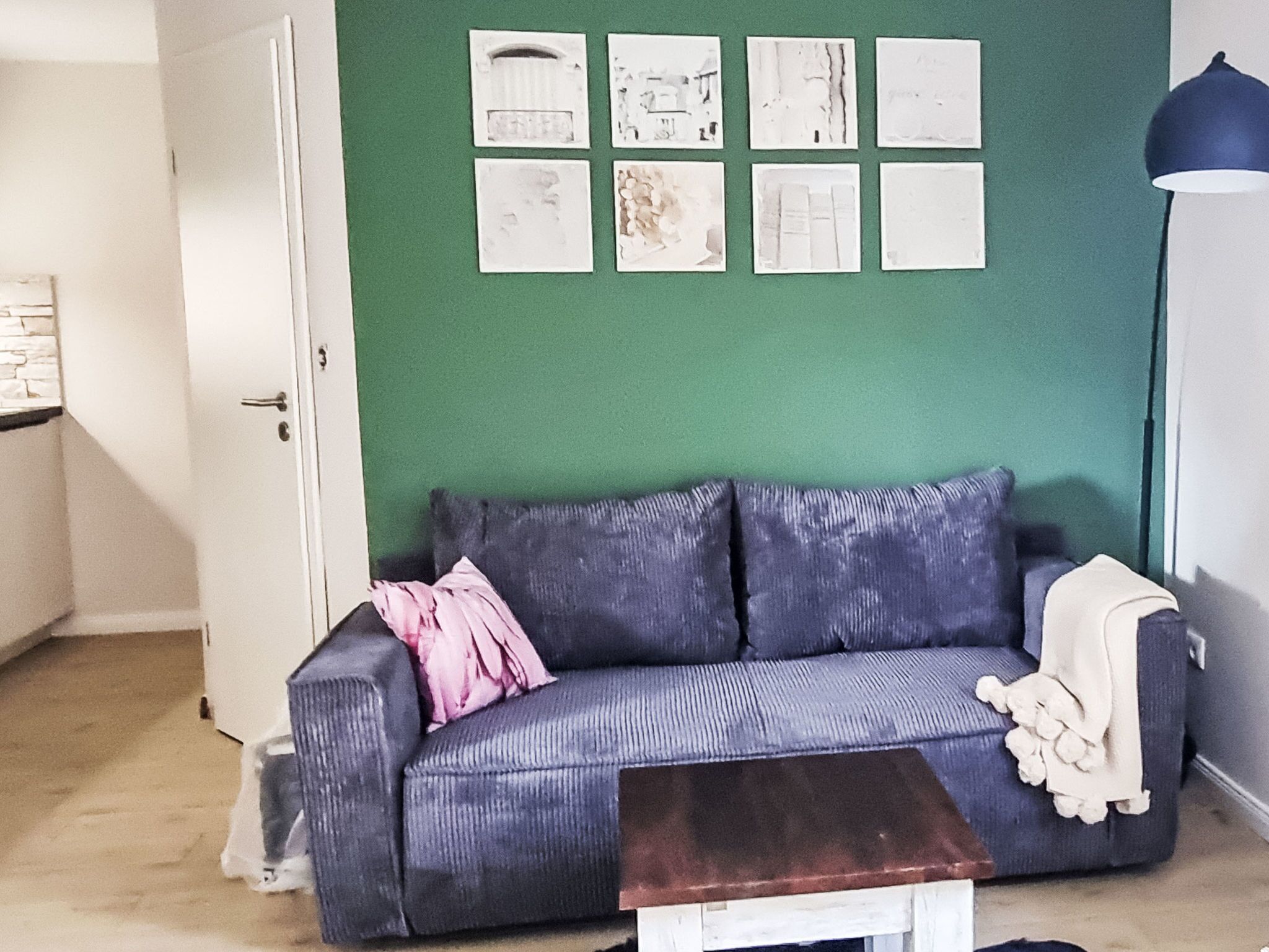 Huoneessa on vihreä seinä, harmaa sohva, jossa on vaaleanpunainen koristetyyny, valkoinen ovi ja pieni valkoinen pöytä.