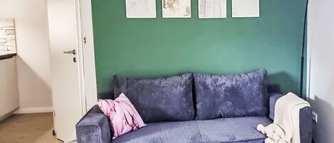 Une pièce avec un mur vert, un canapé gris agrémenté d’un coussin rose, une porte blanche et une petite table blanche.
