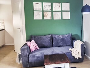 Ein Zimmer mit grüner Wand, einem grauen Sofa mit einem rosafarbenen Kissen, einer weißen Tür und einem kleinen weißen Tisch.