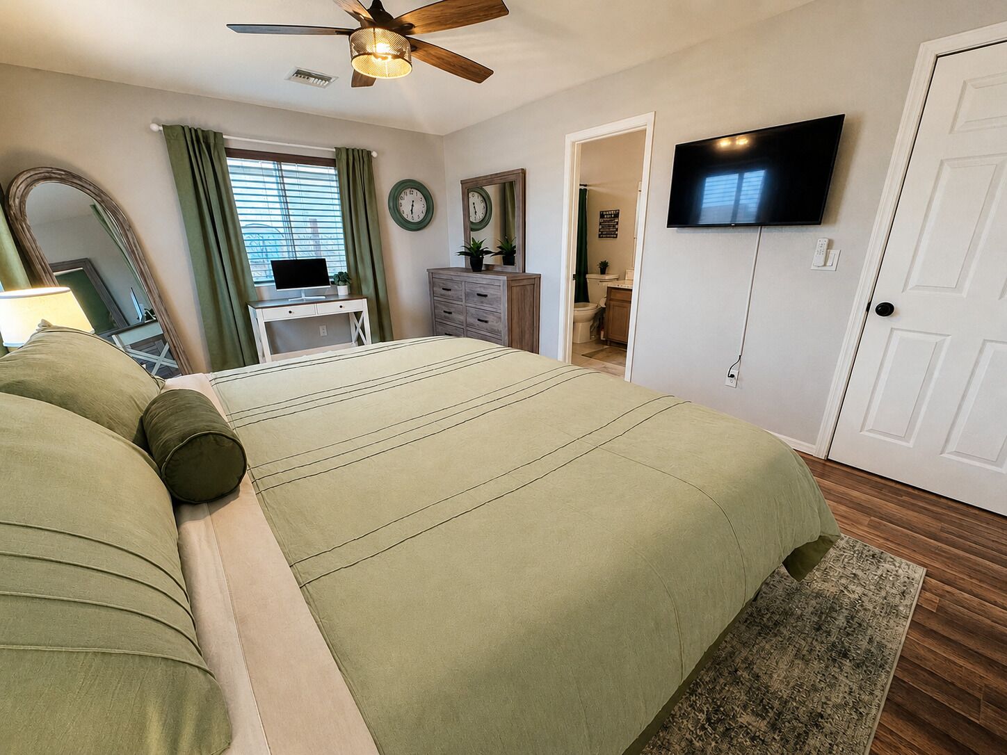 Ein Schlafzimmer mit Bett, Fernseher, Deckenventilator, Schreibtisch und einem durchgehenden Türspalt sichtbaren Badezimmer.