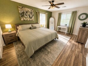 A bedroom with a bed, nightstand, lamp, ceiling fan, artwork, desk, chair, clock, and a potted plant. - Route 66 River House: 8 Min to Boat Launch, Boat & Trailer Parking (Mohave Valley)