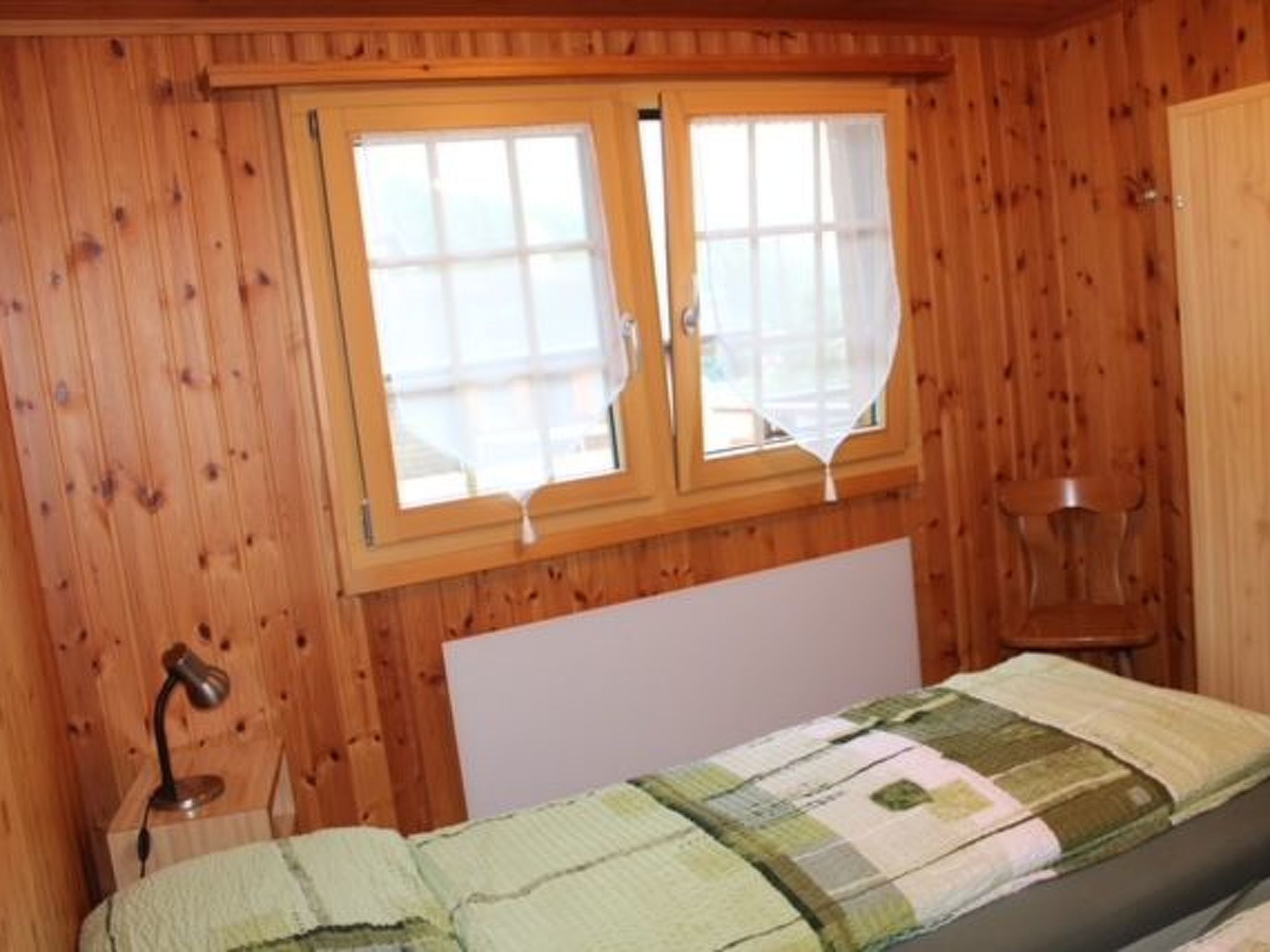 Ein Zimmer mit Holzwänden, einem Bett, einer Nachttischlampe und einem fenster mit Vorhängen.