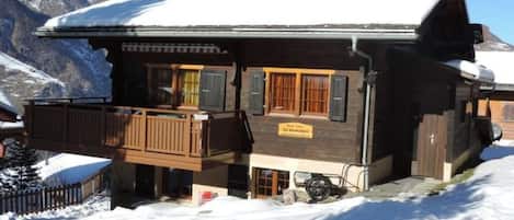 A wooden chalet with a balcony, set against a snowy mountain backdrop.