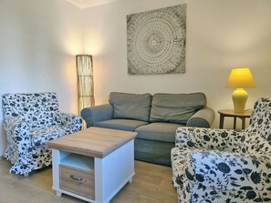 Un salon avec un canapé gris, des fauteuils à motifs floraux, une table basse en bois et une lampe fixée au mur.