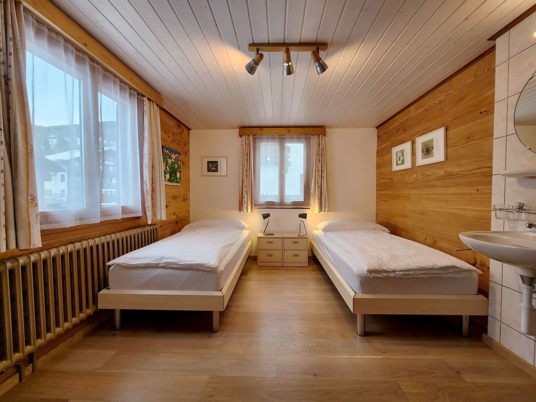 A room with two beds, a window with curtains, a radiator, a wooden wall, and a ceiling light fixture.