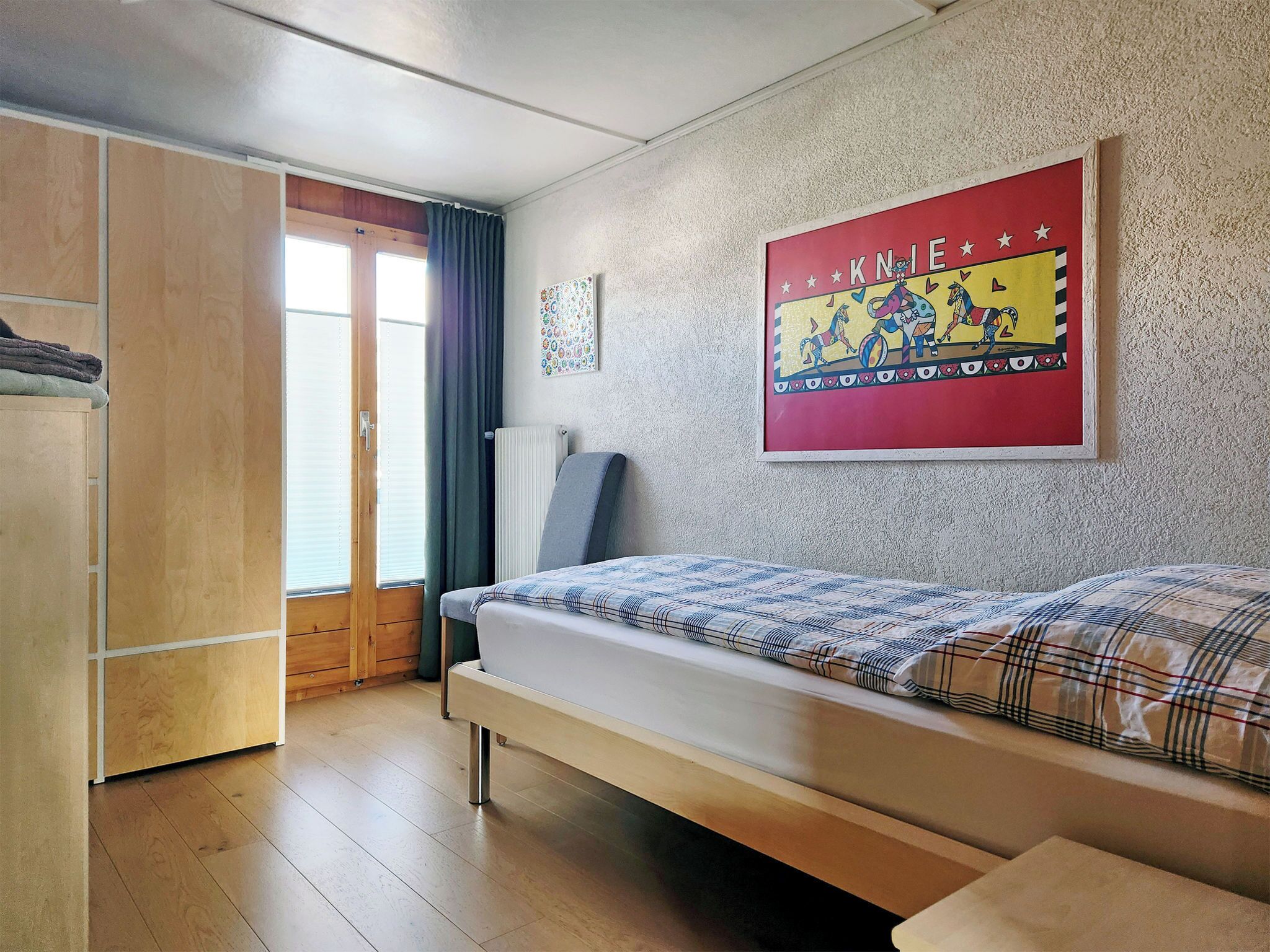 A bedroom with a bed, a wardrobe, a radiator, a window with curtains, and a wall with a colorful poster.