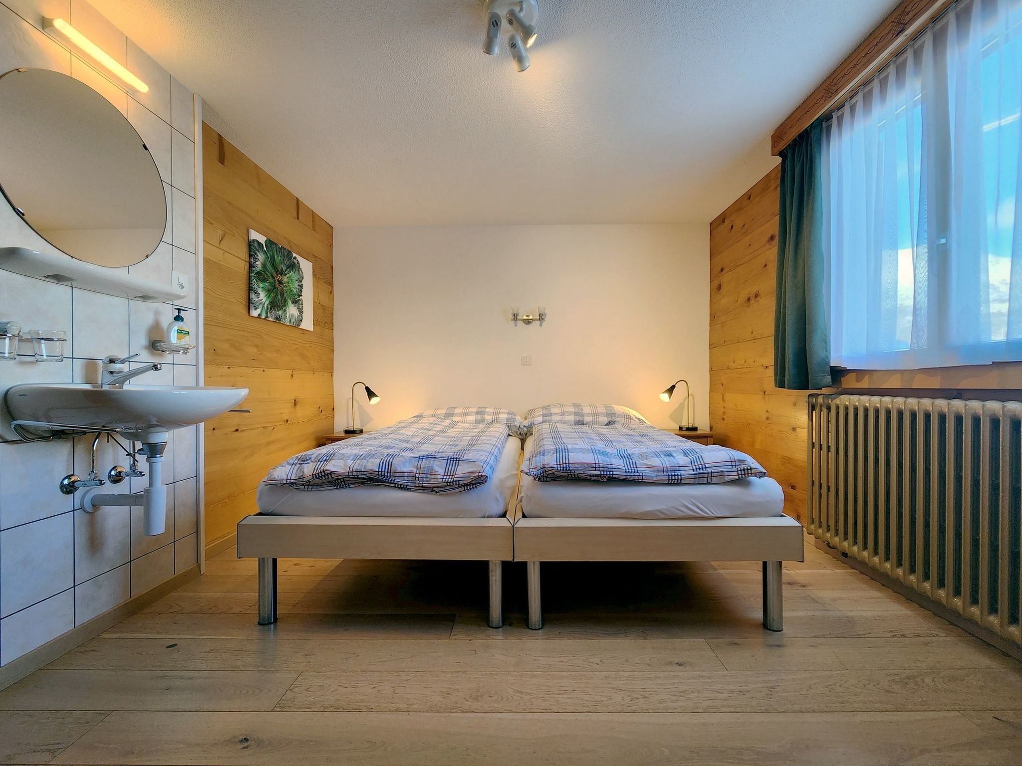 Une chambre à coucher avec un lit double, une table de chevet, un miroir, une fenêtre avec des rideaux et un radiateur.