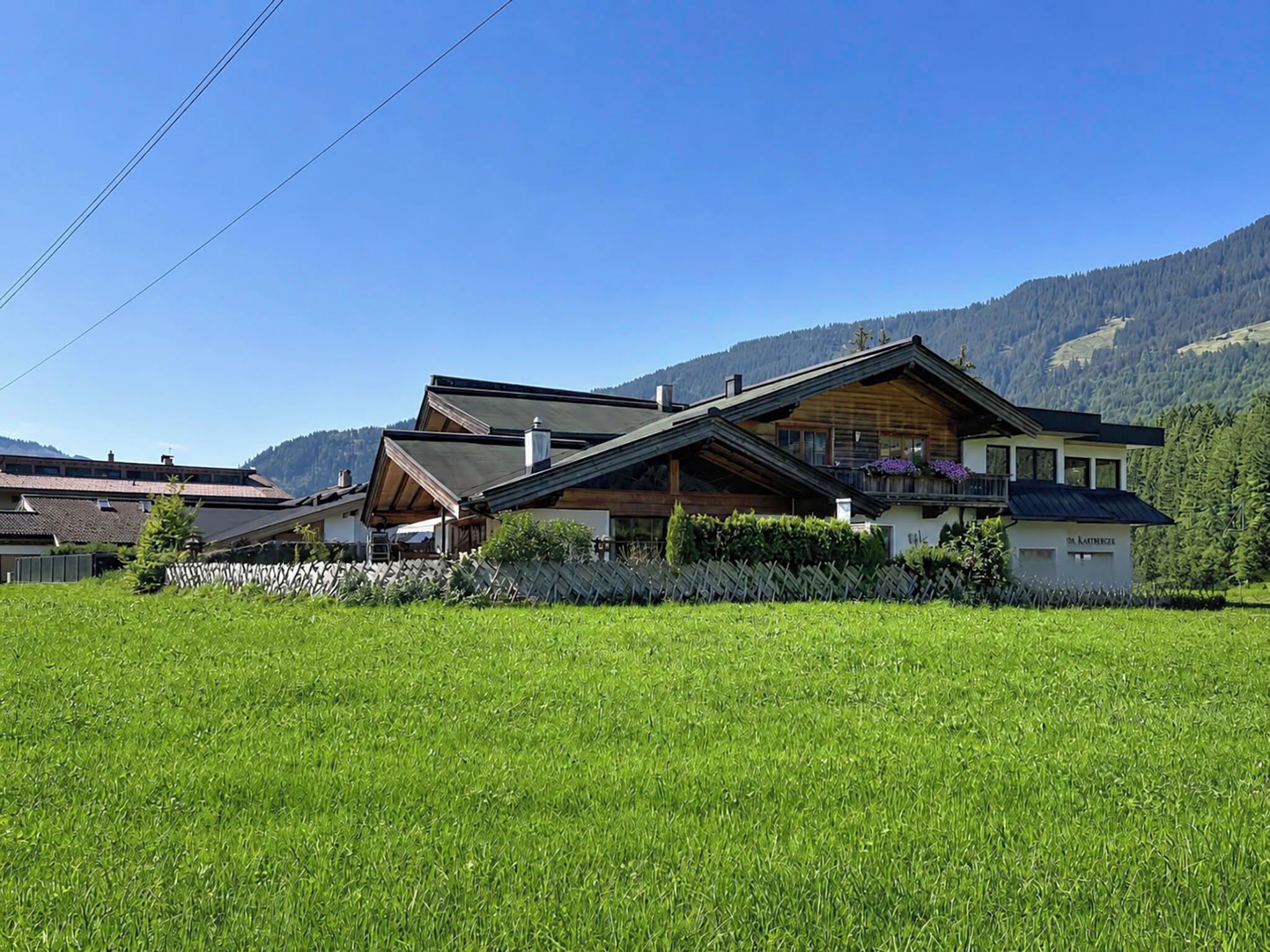 A two-story wooden house with a balcony, surrounded by a well-maintained lawn and a mountain backdrop.