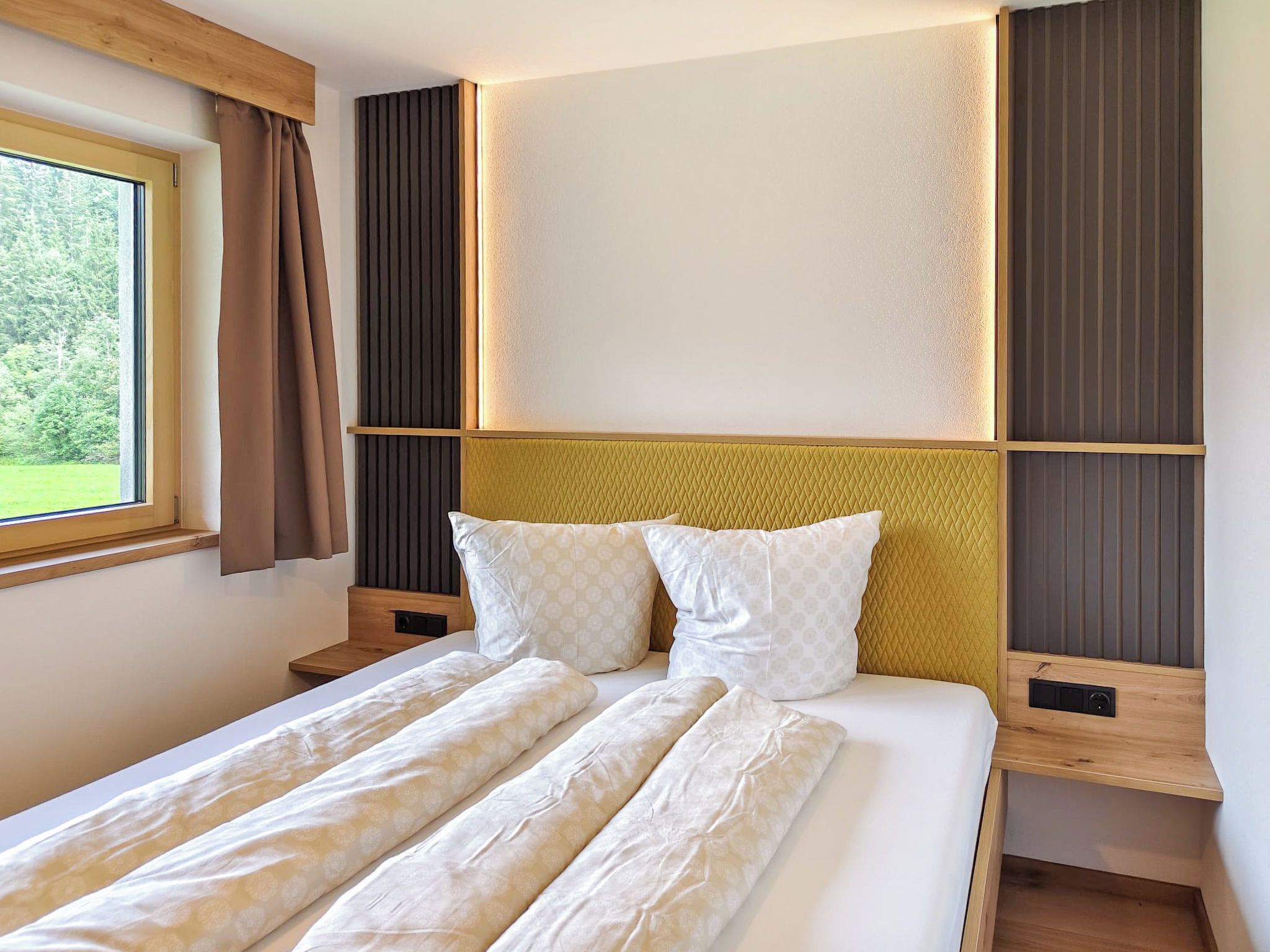 A modern bedroom with a large bed, a window with curtains, and a view of greenery outside.