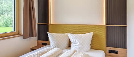 A modern bedroom with a large bed, a window with curtains, and a view of greenery outside.