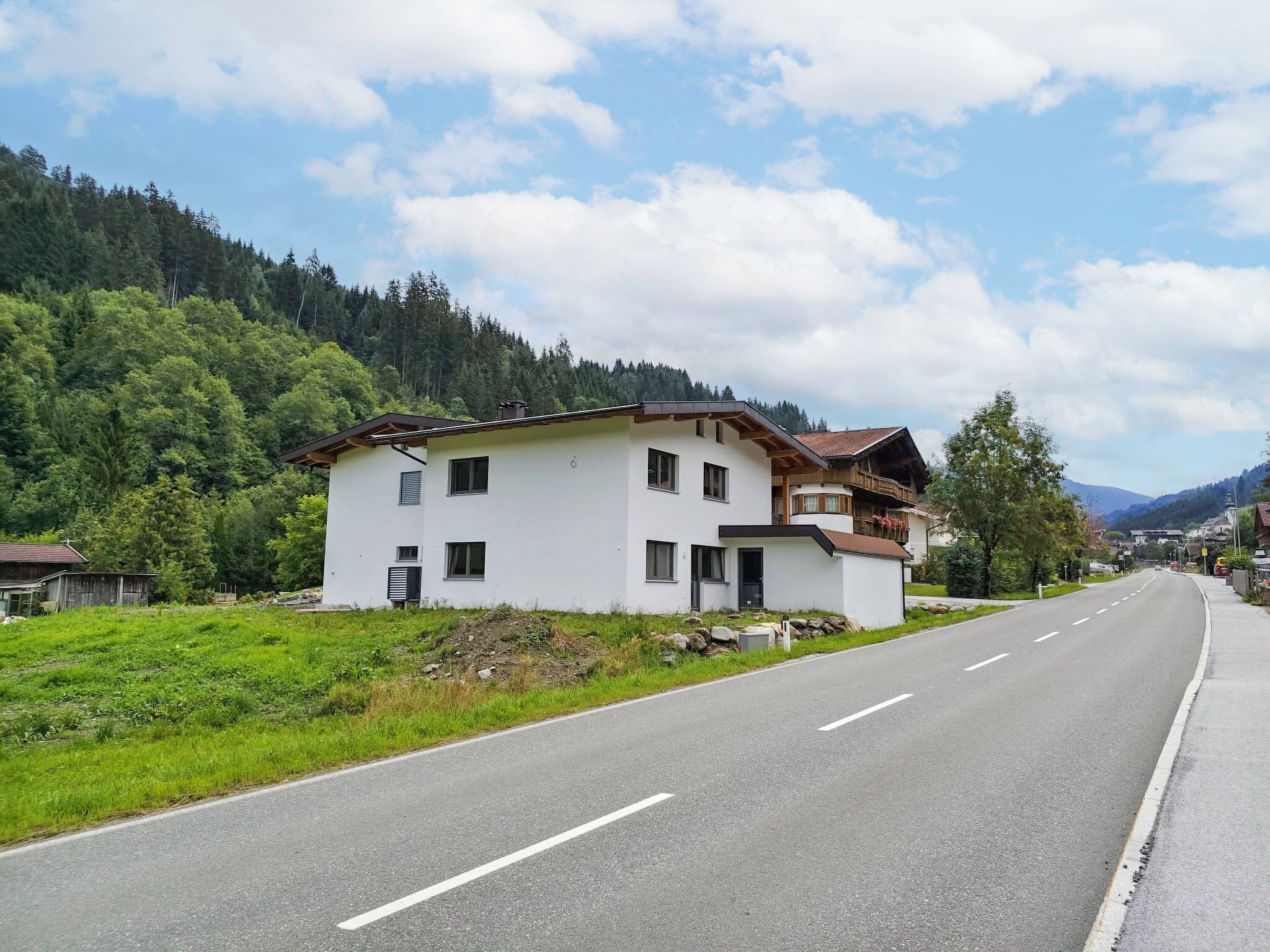 A two-story white building with a balcony, situated beside a road with a forested mountain backdrop.