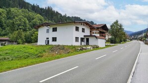 Un bâtiment blanc à deux étages, doté d’un balcon, situé au bord d’une route, avec une montagne boisée en toile de fond.