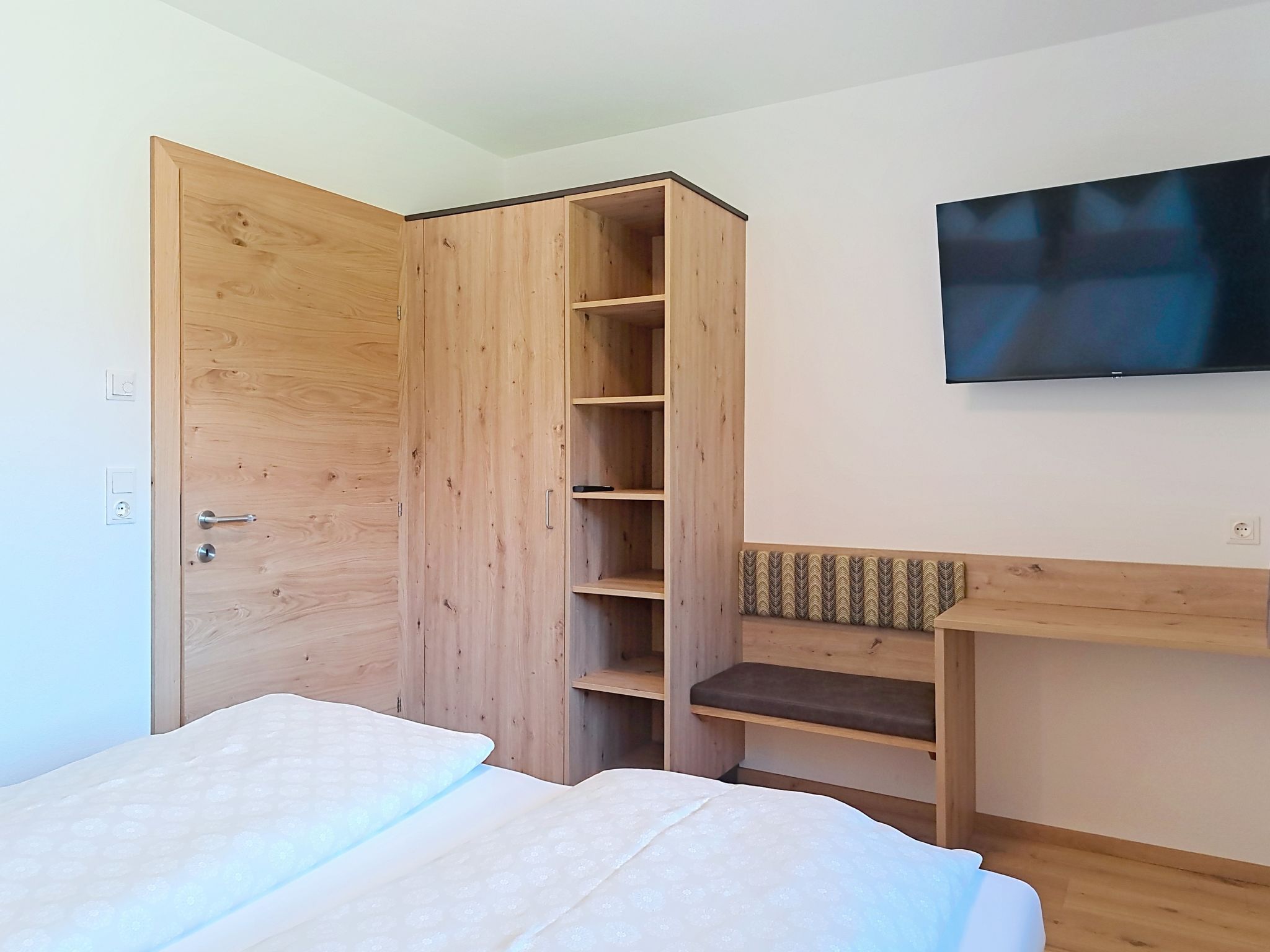 Een slaapkamer met een bed, een houten kledingkast, een bankje en een televisie aan de muur.