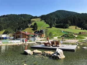 A lakeside resort with clear waters, wooden docks, and a view of a golf course and forested hills in the background. - Apartment Tyrol by Interhome (Auffach)