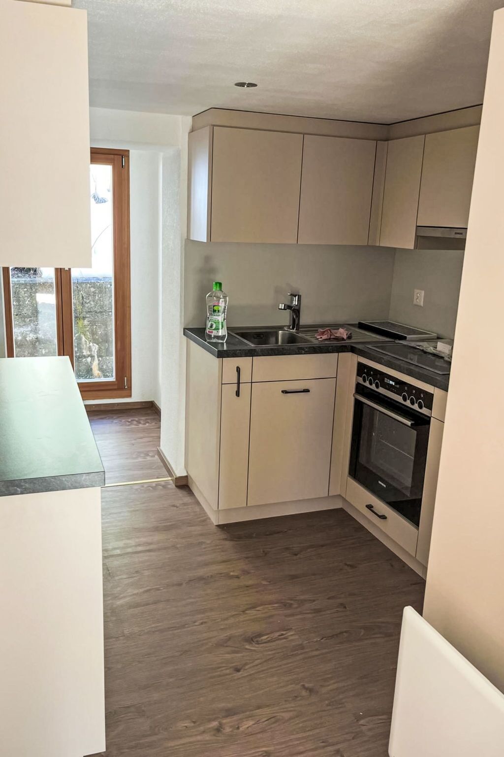 A compact kitchen with a built-in oven, a sink, and a window overlooking a balcony.