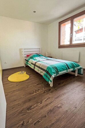 Ein kleines Zimmer mit einem Einzelbett, Holzboden, einem Fenster mit Aussicht und einer gelben Matte.