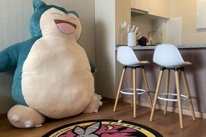 Un jouet en peluche dans une pièce qui comprend un coin cuisine, deux chaises blanches et un plancher en bois.