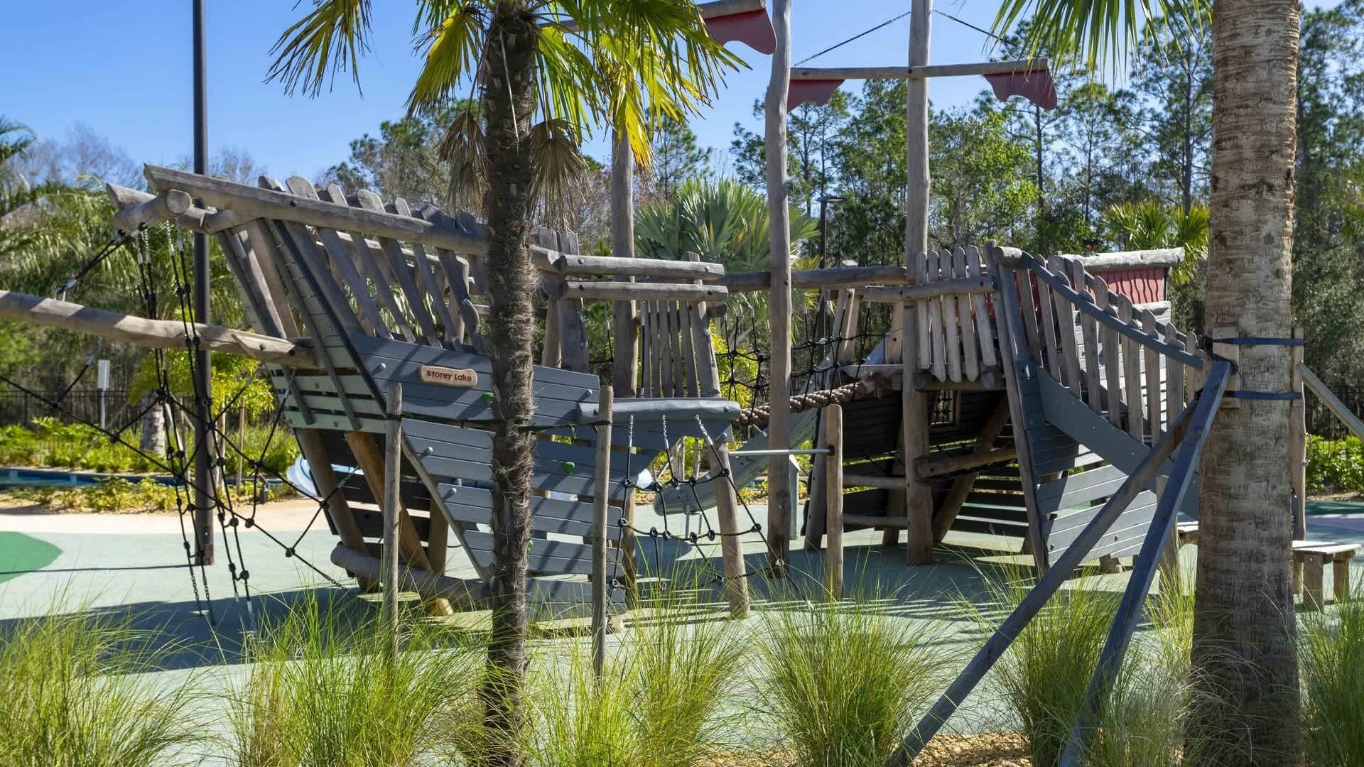 A wooden play structure with ropes and platforms, surrounded by palm trees and greenery.