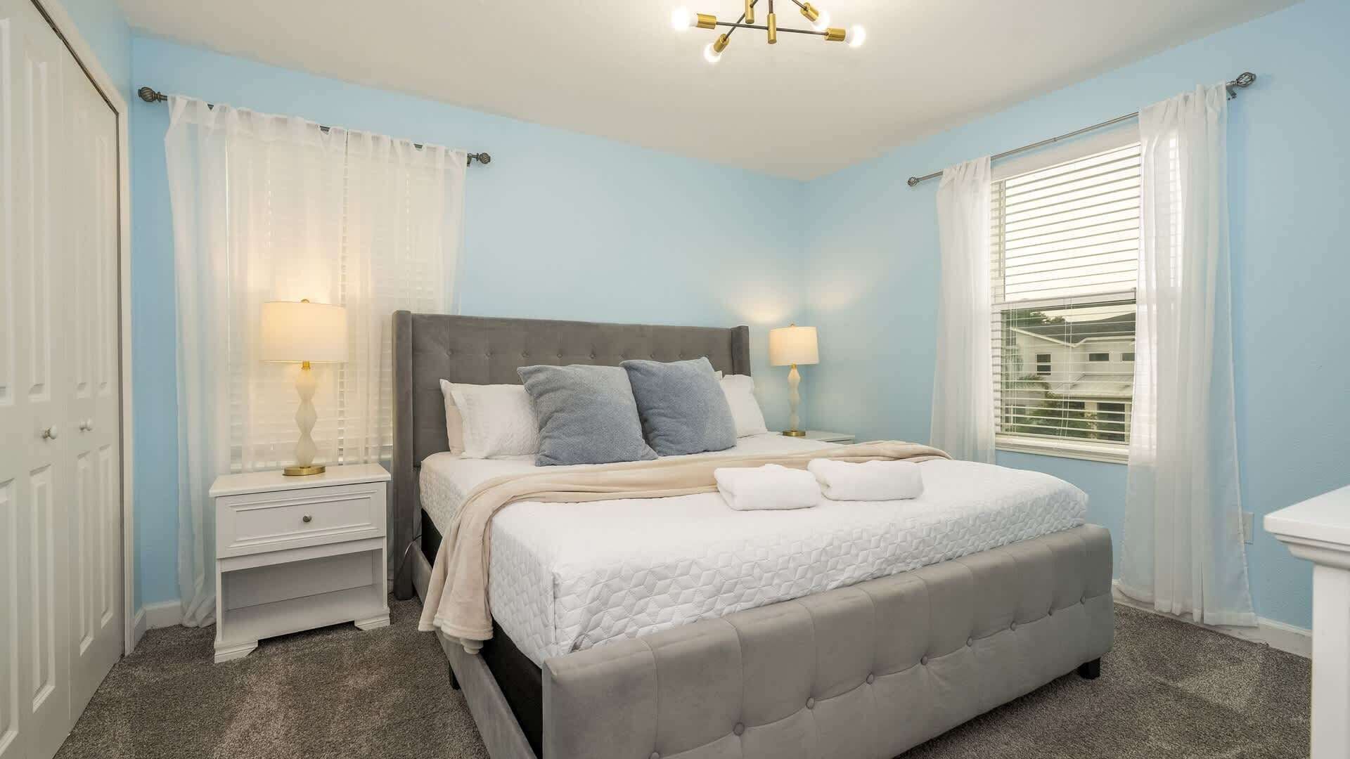 A bedroom with a bed, two bedside tables, a window with blinds, and a chandelier.