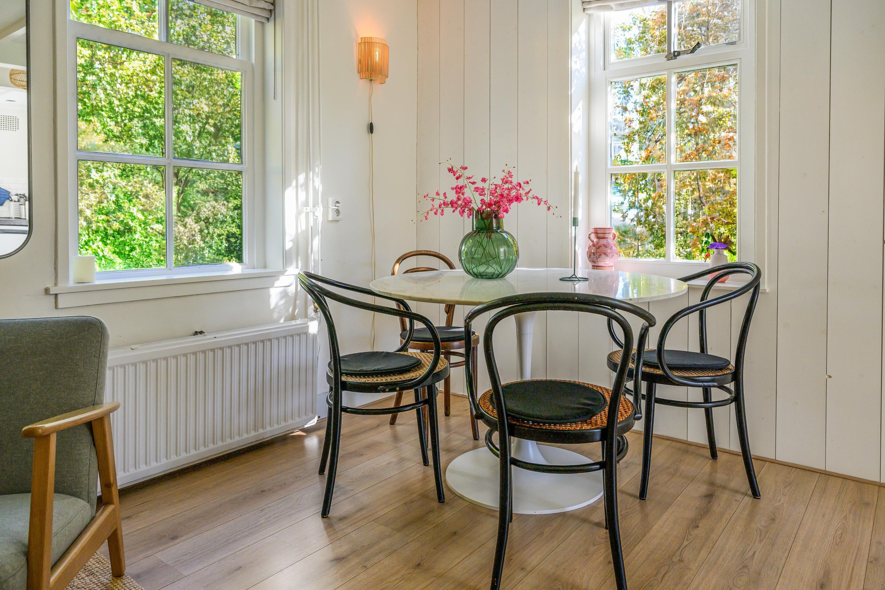 Ein Essbereich mit einem runden Tisch, vier Stühlen, einer Blumenvase und einem Fenster mit Blick auf Bäume.