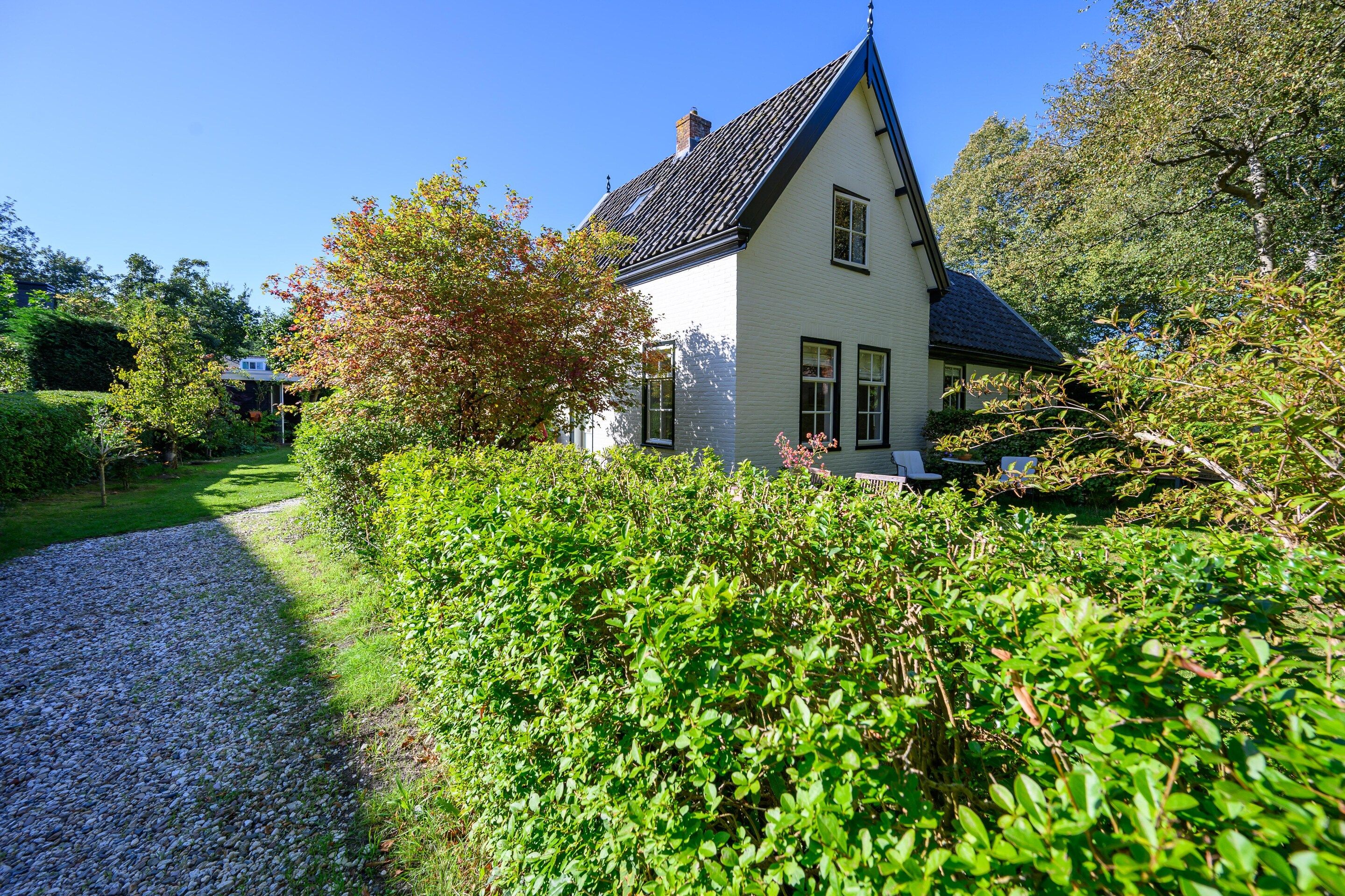 A single-story house with a steeply pitched roof, surrounded by dense shrubbery and a gravel path leading to the entrance.