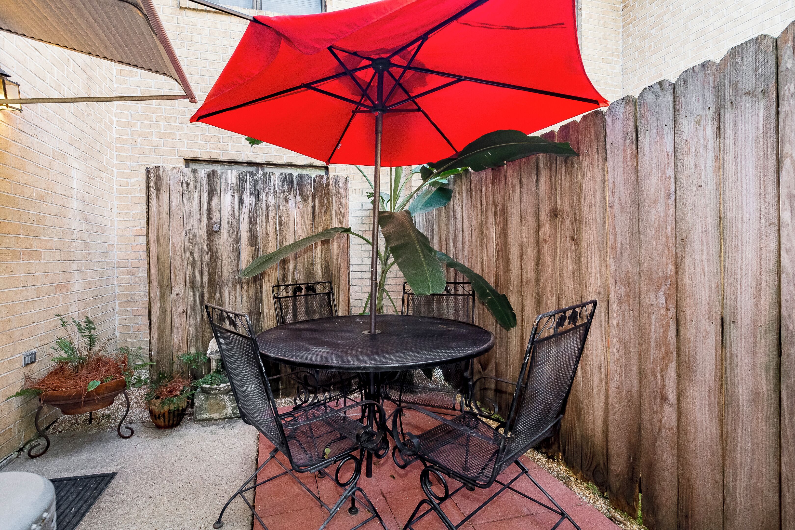 A patio with a round table and chairs, a red umbrella, and a wooden fence.