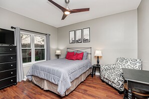Une chambre à coucher comprenant un lit, un bureau, une chaise, un ventilateur de plafond et une fenêtre avec des rideaux.