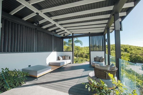 A modern outdoor deck with a wooden floor, a white sofa, a wicker chair, and a glass railing overlooking a lush landscape. - A Stay Worth Traveling For! Tropical Escape with Private Pool and Antigua Views! (Urlings)