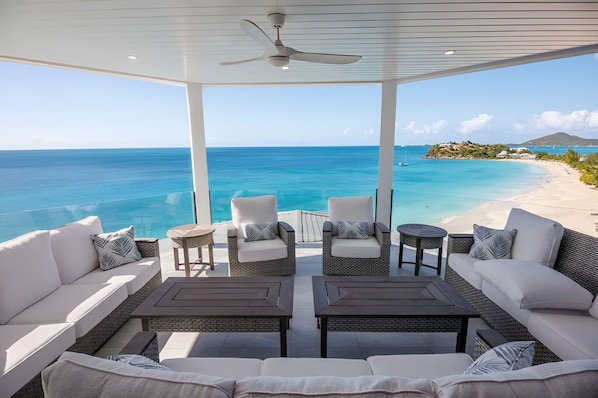 A covered outdoor seating area with wicker furniture, a ceiling fan, and a view of a beach and ocean. - Enjoy Every Moment in a Space Made for Great Getaways! 4BR Villa w/ Private Pool (Urlings)