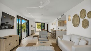 A modern living room with a sofa, armchair, and coffee table. There is a dining area with a table and chairs. The room has large windows and a ceiling fan. - Ocean 20: Luxury 3-Bedroom Oceanfront Suite with Pool & Guest Cottage, Antigua (Urlings)