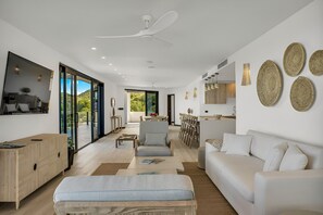 Una sala moderna con sofá, sillón y mesa de centro. Hay un comedor con mesa y sillas. La habitación tiene ventanas amplias y un ventilador de techo.