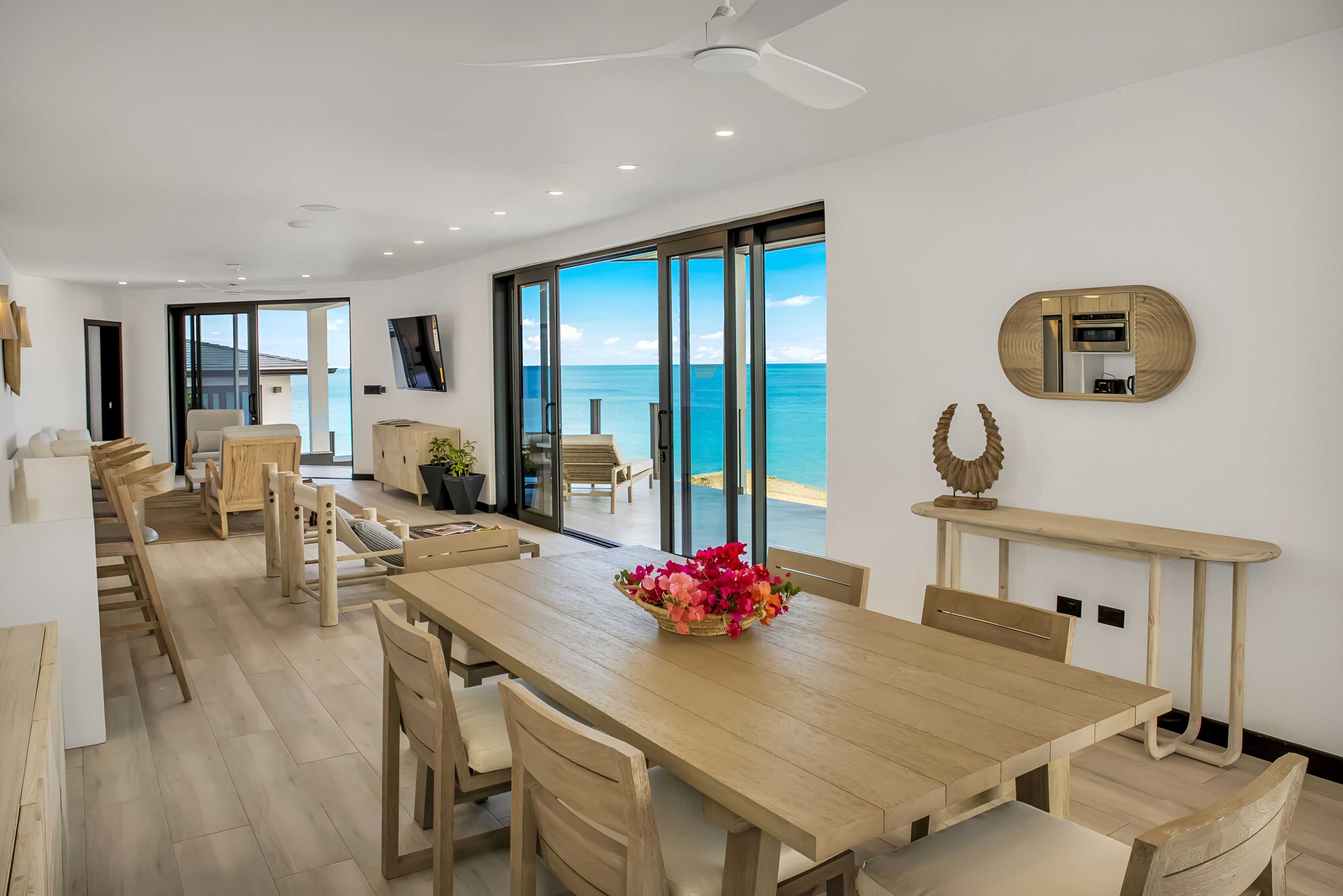 A modern dining area with a wooden table, chairs, and a large window offering a view of the ocean.