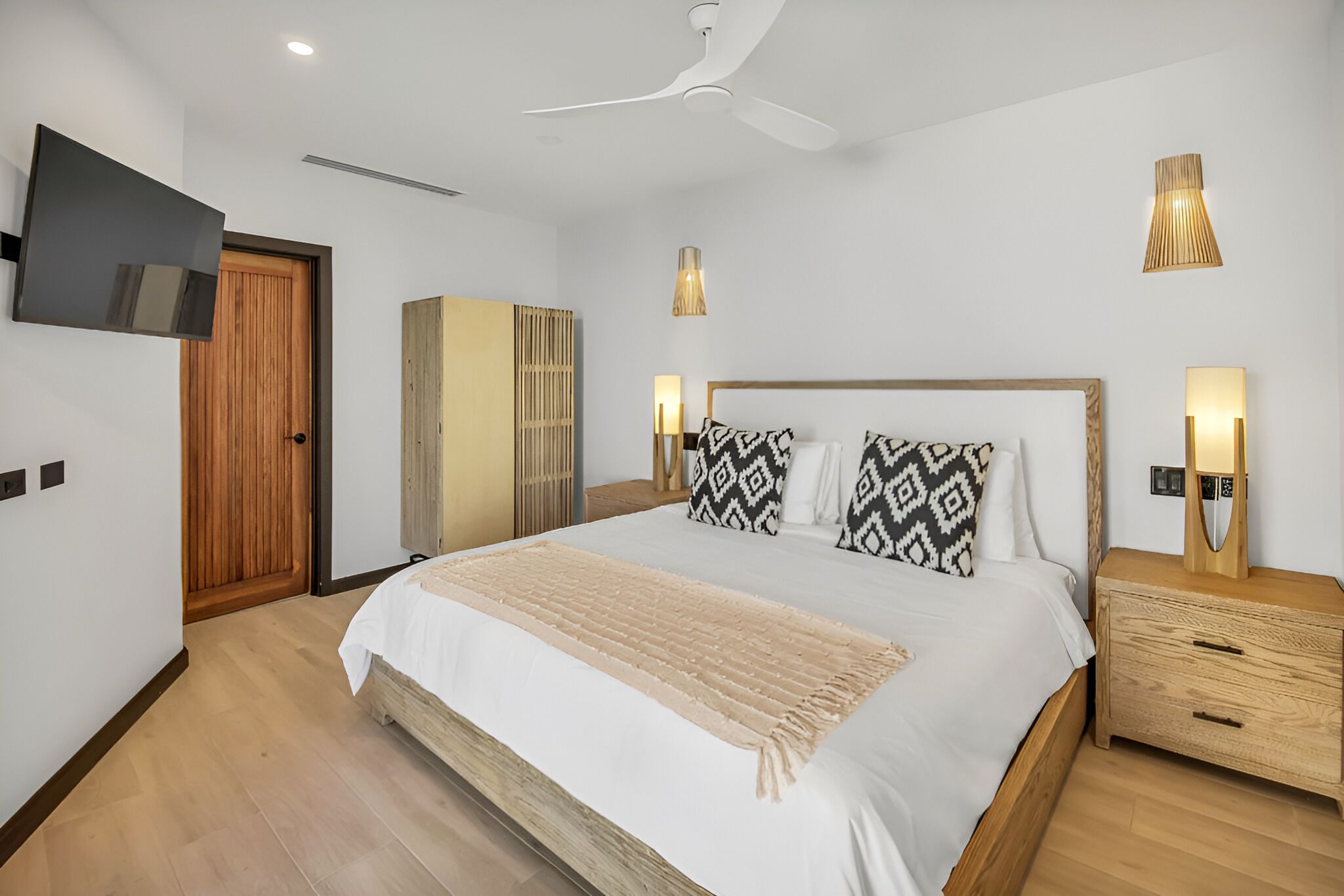 A modern bedroom with a large bed, wooden headboard, bedside lamps, a ceiling fan, and a TV mounted on the wall.