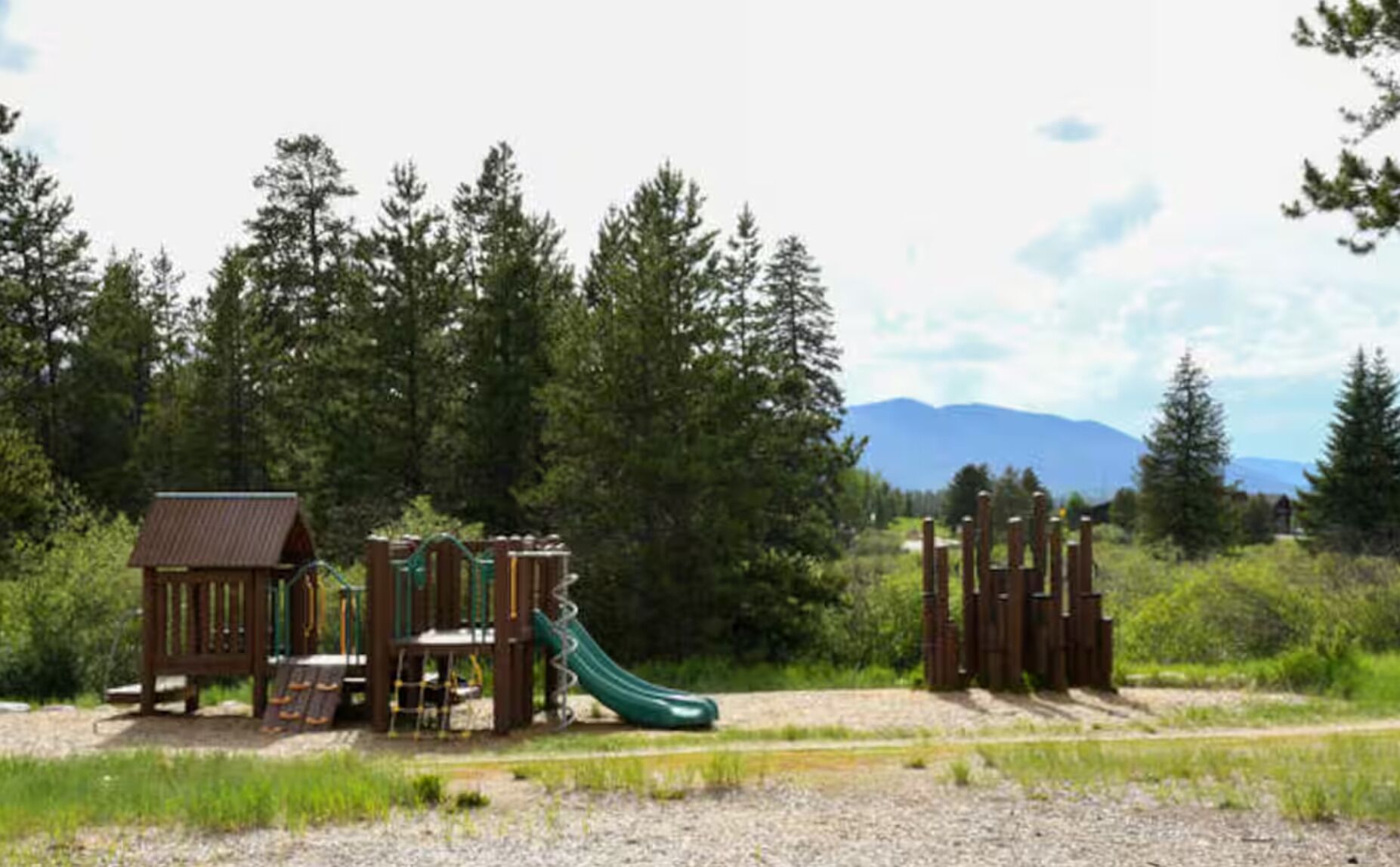 A playground with a slide and climbing structures, surrounded by trees and a mountain in the background.