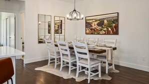 A dining room with a white table set for six, white chairs, a chandelier, and a large landscape painting on the wall. - Luxury 3 Bed Townhome • Walk to Town • on Ski Bus • Hot Tub • 2-Car Garage (Fraser)