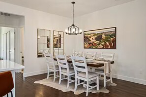 A dining room with a white table set for six, white chairs, a chandelier, and a large landscape painting on the wall. - Luxury 3 Bed Townhome • Walk to Town • on Ski Bus • Hot Tub • 2-Car Garage (Fraser)
