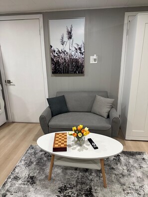 A living room with a grey sofa, a white coffee table with a chessboard and a vase of flowers, and a painting on the wall.
