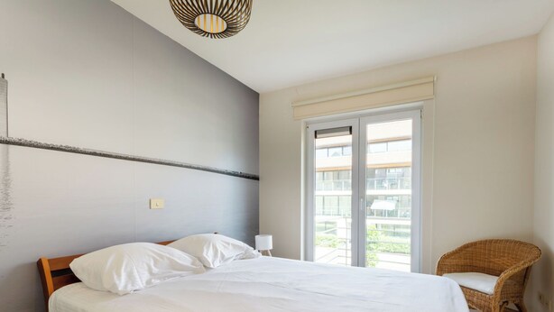 Un dormitorio con una cama grande, una ventana con vistas a edificios, una silla de mimbre y una lámpara fijada a la pared.