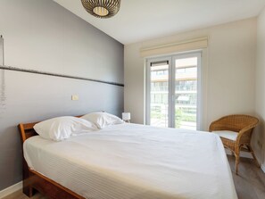 Un dormitorio con una cama grande, una ventana con vistas a edificios, una silla de mimbre y una lámpara fijada a la pared.