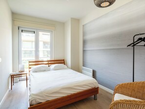 A bedroom with a bed, a bedside table, a lamp, a radiator, and a window with a view of buildings. - Goldsands III with parking by Interhome (De Haan)