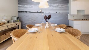 Un comedor con una mesa de madera dispuesta para seis personas, rodeado de sillas de mimbre, un cuadro grande de una playa y una lámpara colgante moderna.