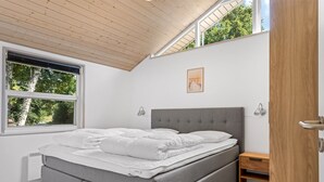 Ein Schlafzimmer mit grauem Polsterbett, einem Holznachttisch, einem Fenster mit Jalousien und einer Holzbalkendecke.
