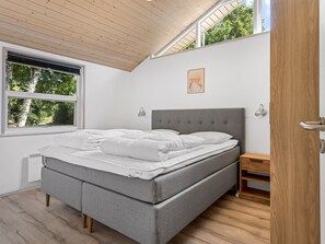 Een slaapkamer met een grijs gestoffeerd bed, een houten nachtkastje, een raam met jaloezieën en een plafond met houten balken.