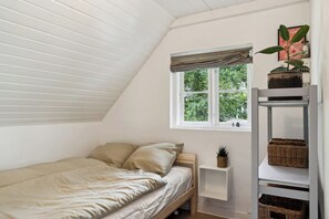 A bedroom with a sloped ceiling, a single bed with a beige duvet, a window with a striped valance, and a shelf with baskets. - Fine old details near Stubbe Lake (Ebeltoft)