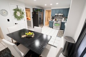 Eine moderne Küche mit Essbereich, ausgestattet mit einem schwarzen Tisch, einer Obstschale, weißen Stühlen und einem Mülleimer aus Edelstahl.